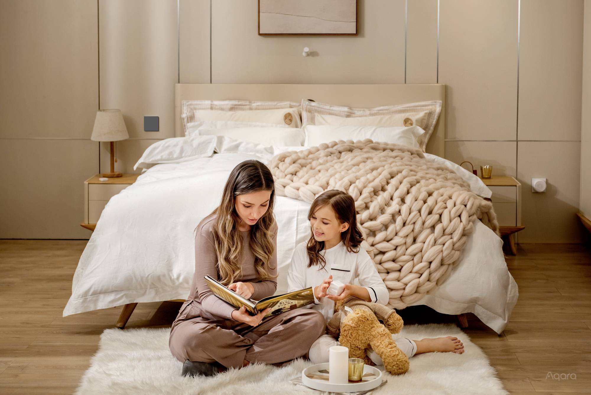 Una donna e una bambina sono sedute su un tappeto davanti a un letto e leggono insieme un libro. Entrambe sembrano rilassate e sorridenti.