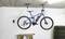 Una bicicletta blu è appesa a un supporto da soffitto in un garage. A destra sono visibili pneumatici impilati, sotto i quali si trova un bancone di legno.