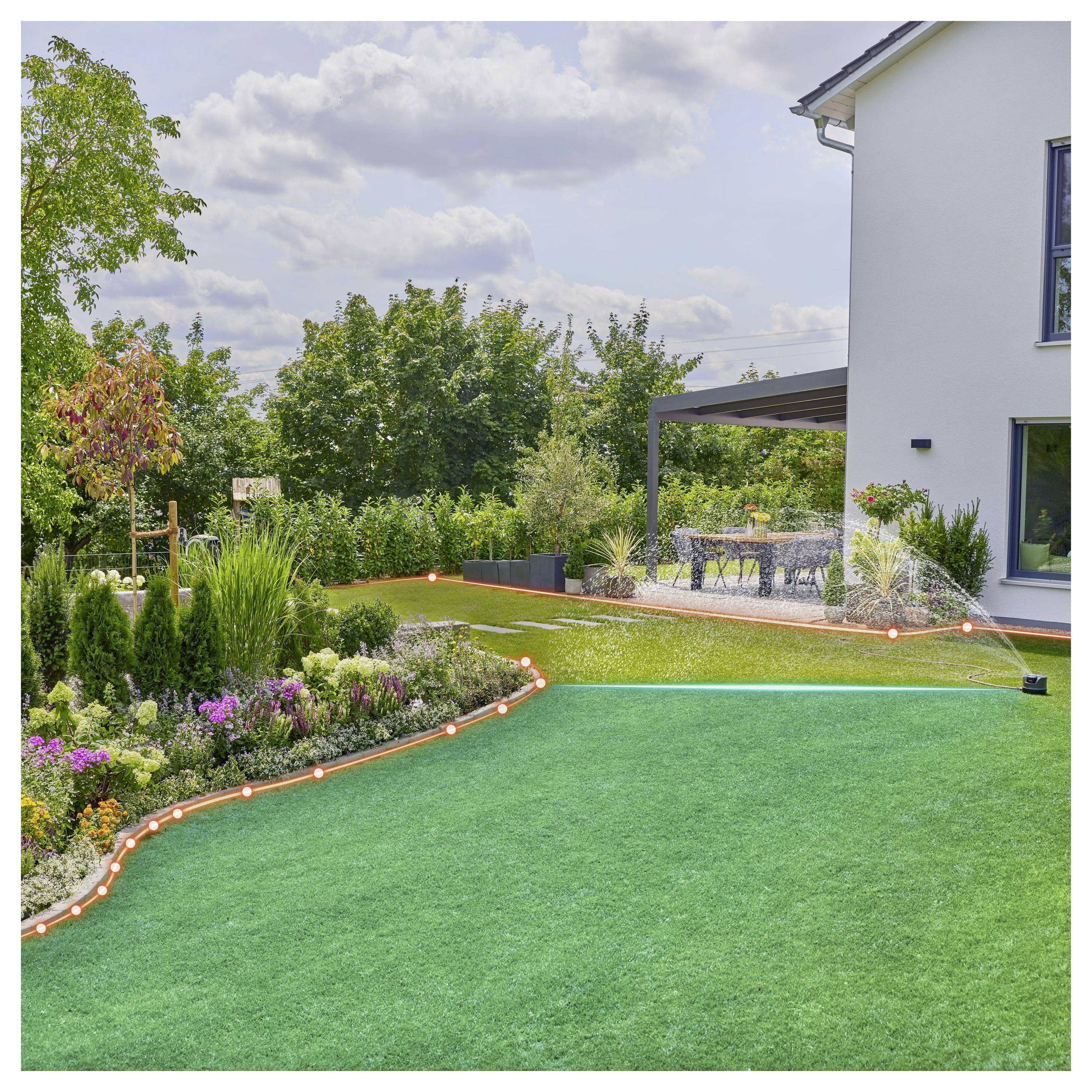 Un rigoglioso giardino nel cortile presenta un prato con aiuole di fiori colorati e un'area patio. Un irrigatore innaffia l'erba sotto un cielo parzialmente nuvoloso.