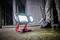 Una lampada da cantiere luminosa illumina un ambiente semibuio. Sullo sfondo si vedono le gambe di una persona vestita con abiti da lavoro.