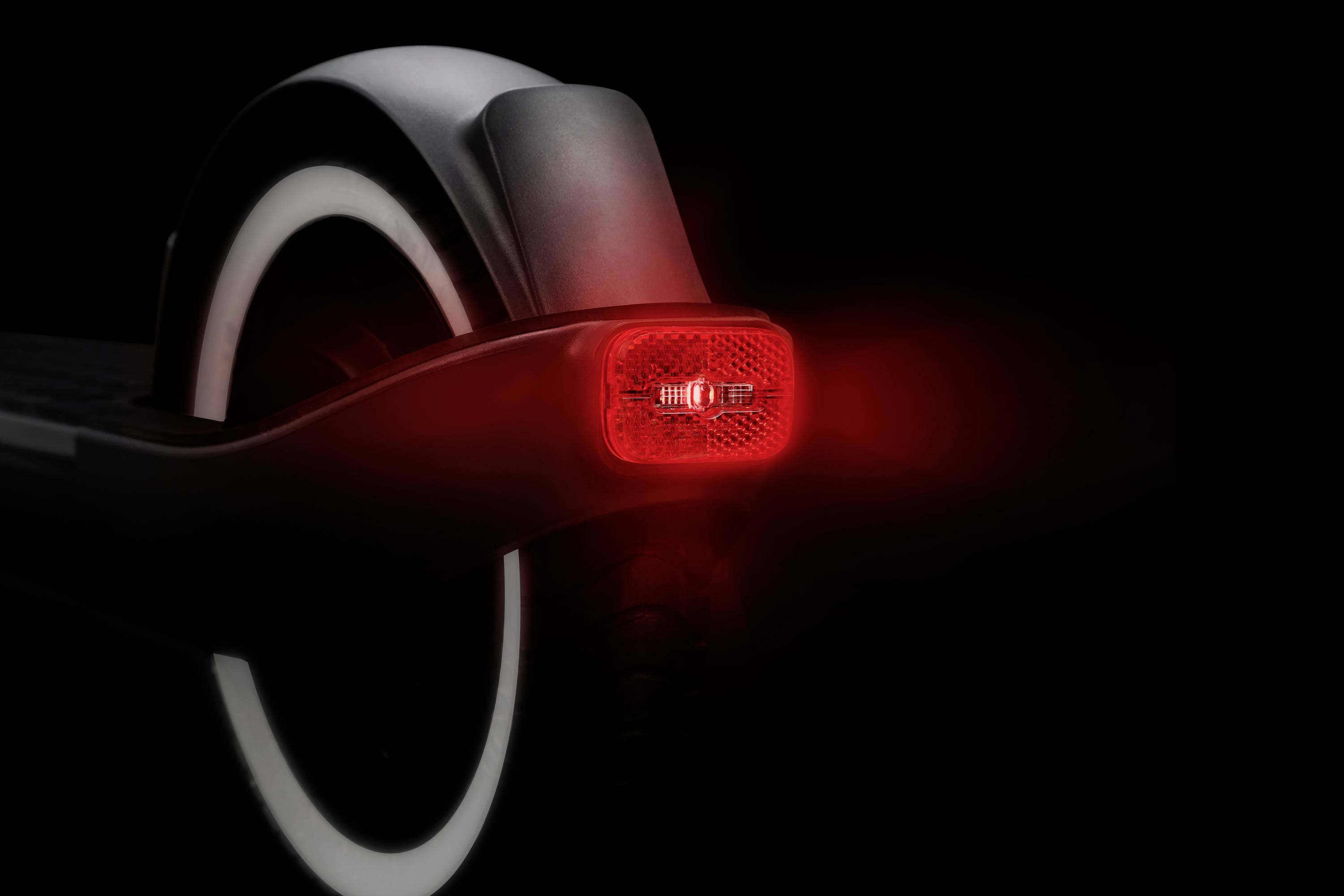 Un monopattino elettrico visto da dietro, con faro posteriore rosso illuminato e superficie riflettente, su sfondo nero.