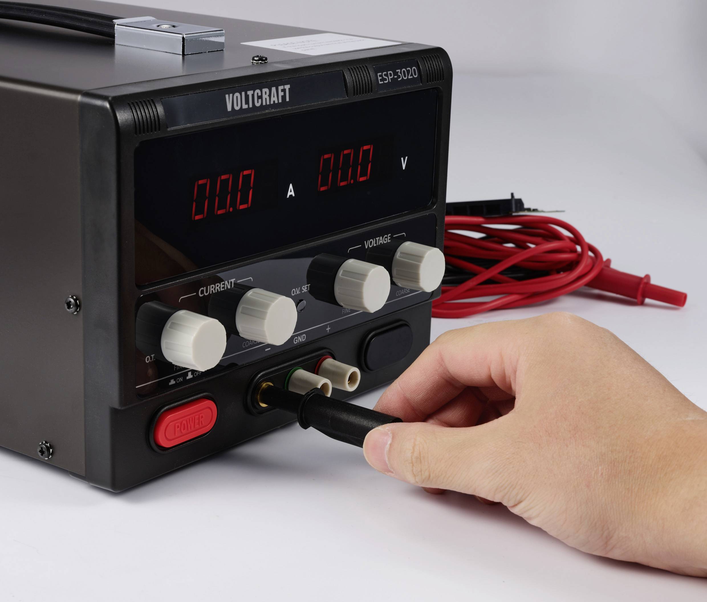 Una mano inserisce un cavo in un'alimentazione Voltcraft ESP-3020. Il display mostra '0.0 V' e '0.0 A'.
