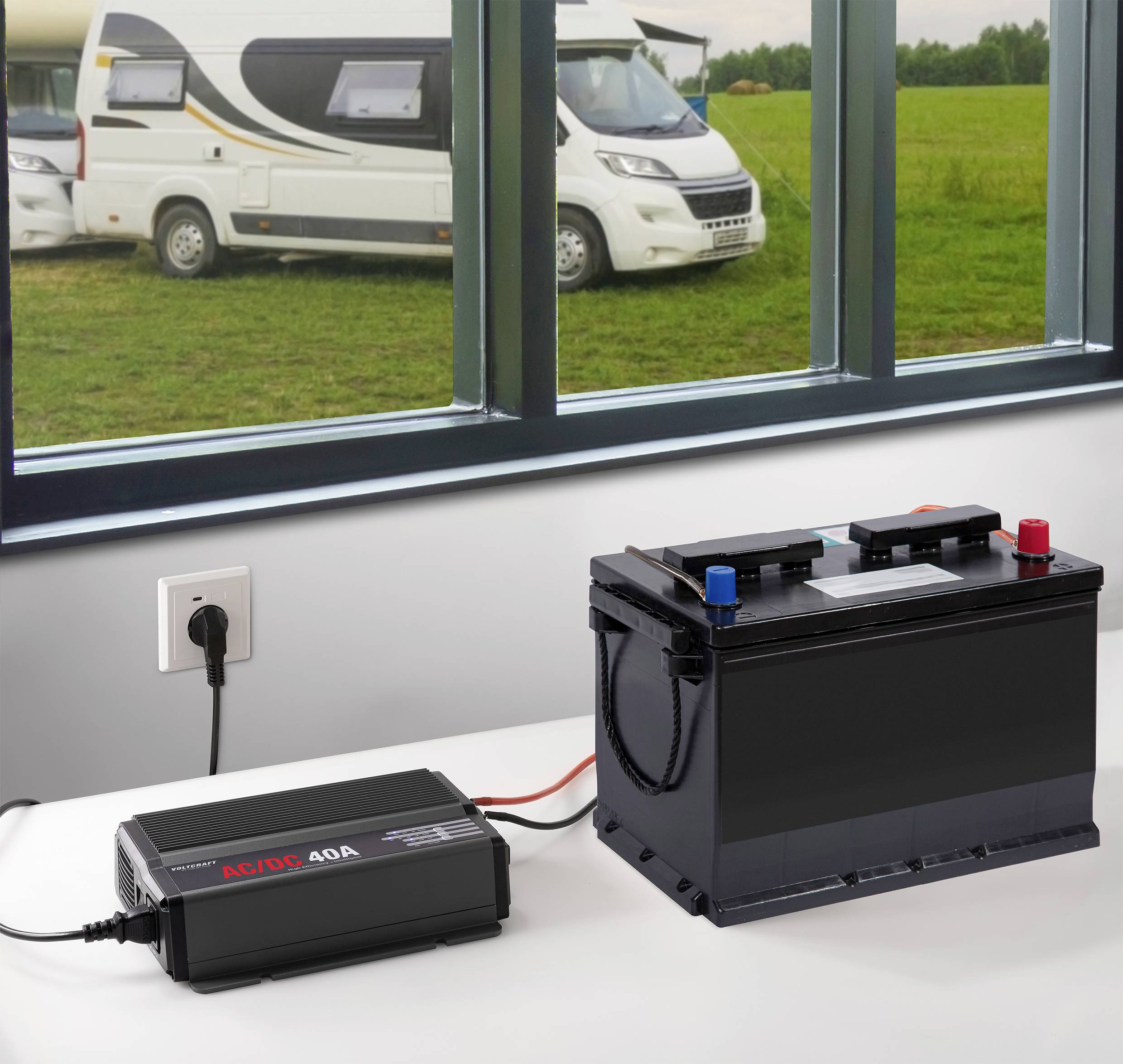 Un caricabatterie è collegato a una batteria per auto su un tavolo. Sullo sfondo sono visibili camper e un campo verde.