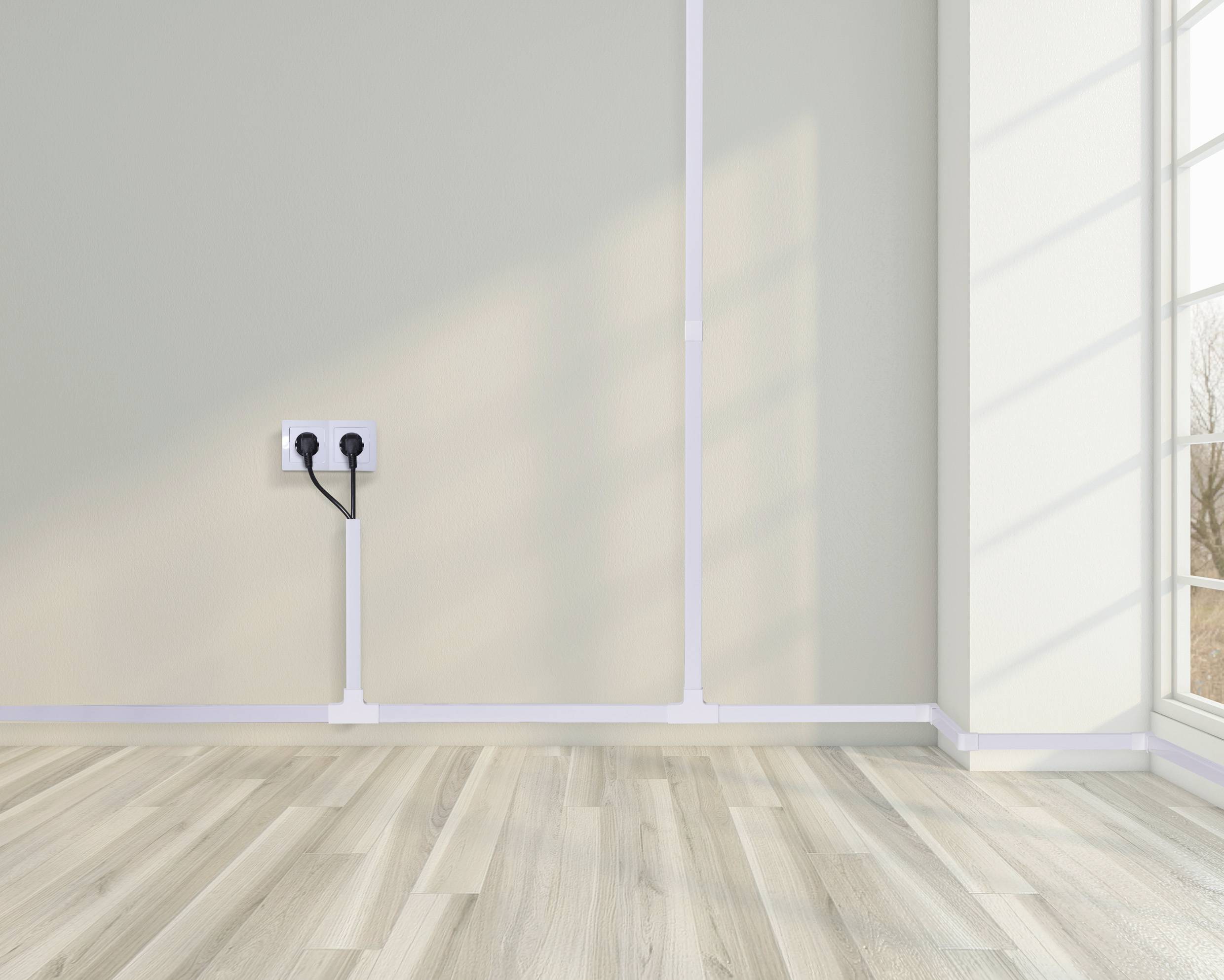 Una stanza vuota con pavimento in legno chiaro, una presa elettrica con due cavi inseriti sulla parete e una grande finestra.