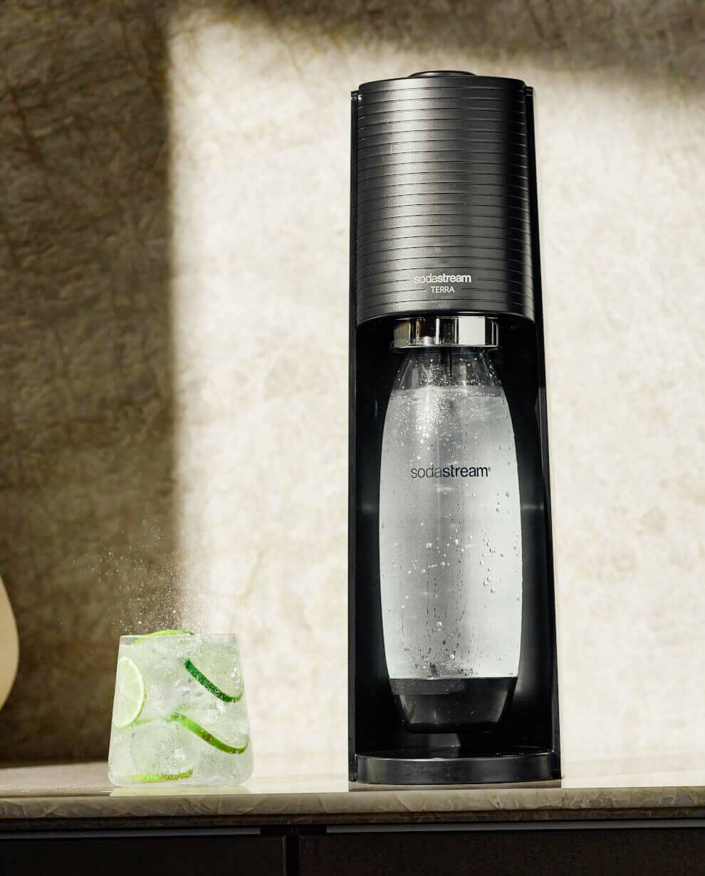 Una macchina Sodastream nera è posizionata su un bancone. Accanto a essa un bicchiere con acqua frizzante e fette di lime.