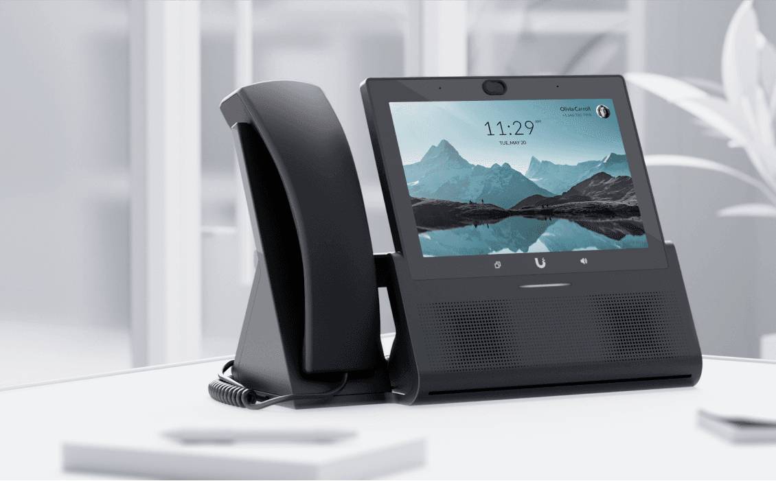 Telefono nero con touchscreen mostra le 11:29 e un paesaggio montano. Posizionato su un tavolo bianco in un ufficio moderno.