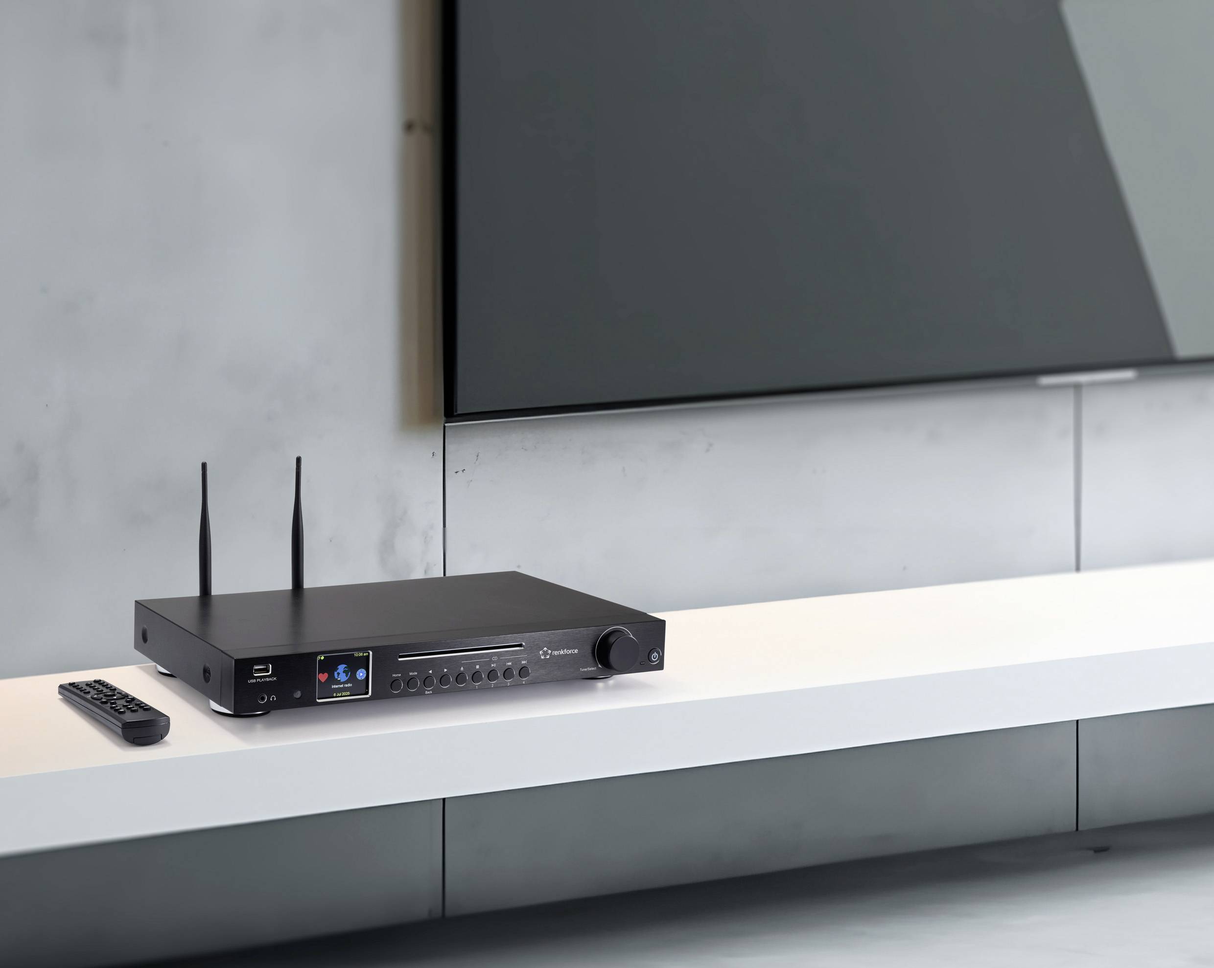 Un ricevitore audio nero con due antenne è posizionato su una mensola bianca sotto una TV a schermo piatto montata a parete, accompagnato da un telecomando.