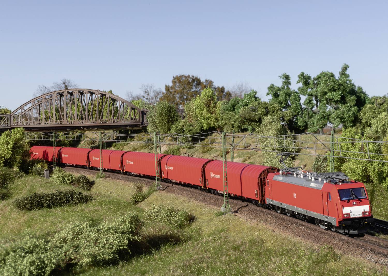 Un treno merci rosso passa sotto un ponte di ferro attraverso un paesaggio verde. Scena di un plastico ferroviario con alberi e ambiente dettagliato.