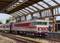 Un modellino di treno con un convoglio rosso e grigio sta viaggiando sotto una tettoia ferroviaria. Alcune persone sono in piedi sul marciapiede.