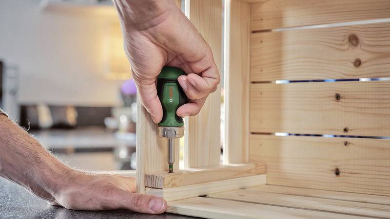Una persona sta assemblando una scaffalatura in legno con un cacciavite. Le mani tengono lo scaffale fermo. Sullo sfondo si intravede una cucina sfocata.