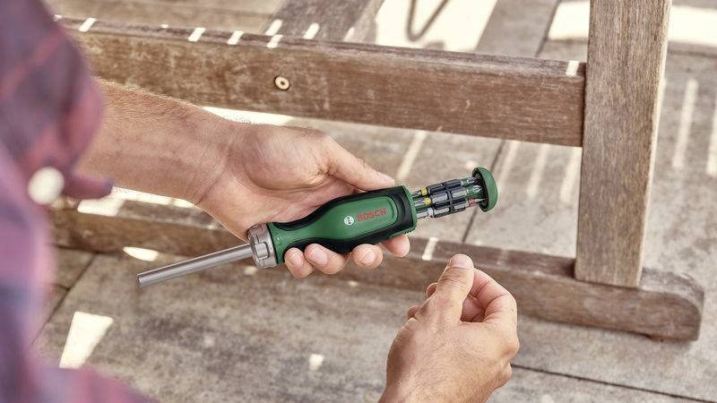 Una persona tiene un cacciavite verde con punte intercambiabili in mano. Sullo sfondo è visibile un tavolo di legno.