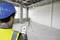 Un lavoratore edile con casco e giubbotto di sicurezza utilizza uno smartphone per controllare uno strumento di misurazione su un treppiede in una stanza vuota.