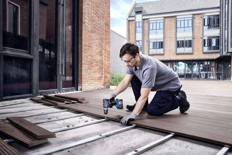 Un uomo lavora all'aperto su una terrazza, sta posando tavole di legno con un trapano a batteria e indossa occhiali protettivi e guanti. Sullo sfondo è visibile un edificio.