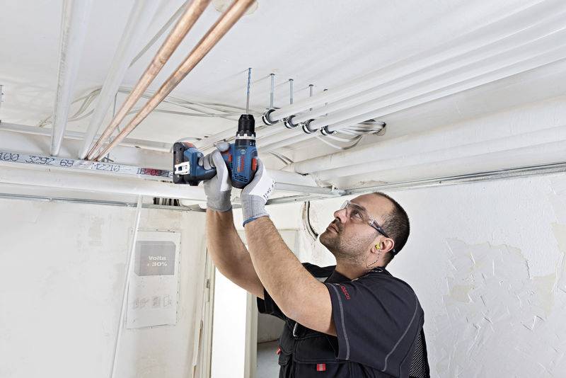 Un artigiano fissa dei tubi al soffitto di una stanza utilizzando un avvitatore a batteria, indossando occhiali protettivi e guanti da lavoro.