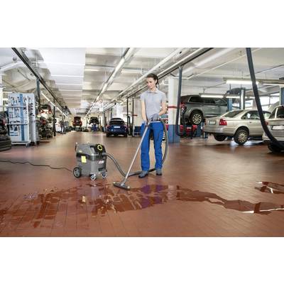Una persona sta pulendo il pavimento di un'officina meccanica con un aspiraliquidi per rimuovere pozze d'acqua. Sullo sfondo sono visibili alcune automobili.