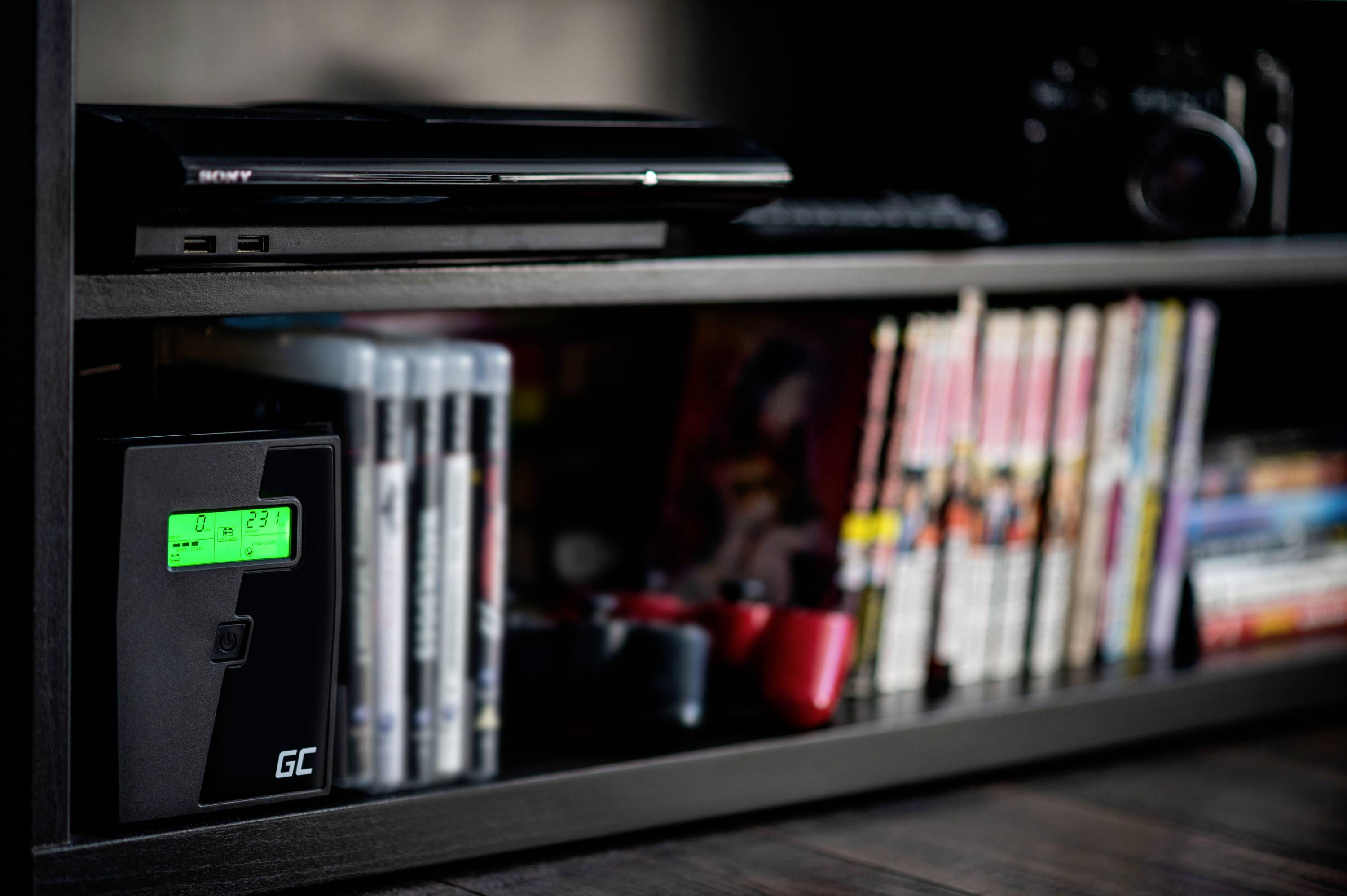 Una scaffalatura nera contiene DVD, giochi, una console per videogiochi e un router. In primo piano è visibile un dispositivo con un display illuminato.