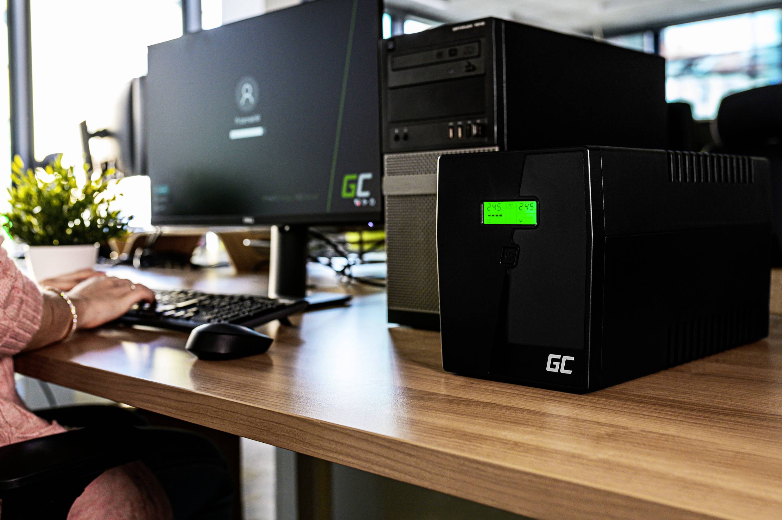 Un posto di lavoro con computer, schermo, una persona seduta alla scrivania e un'unità di alimentazione di continuità (UPS) in primo piano.