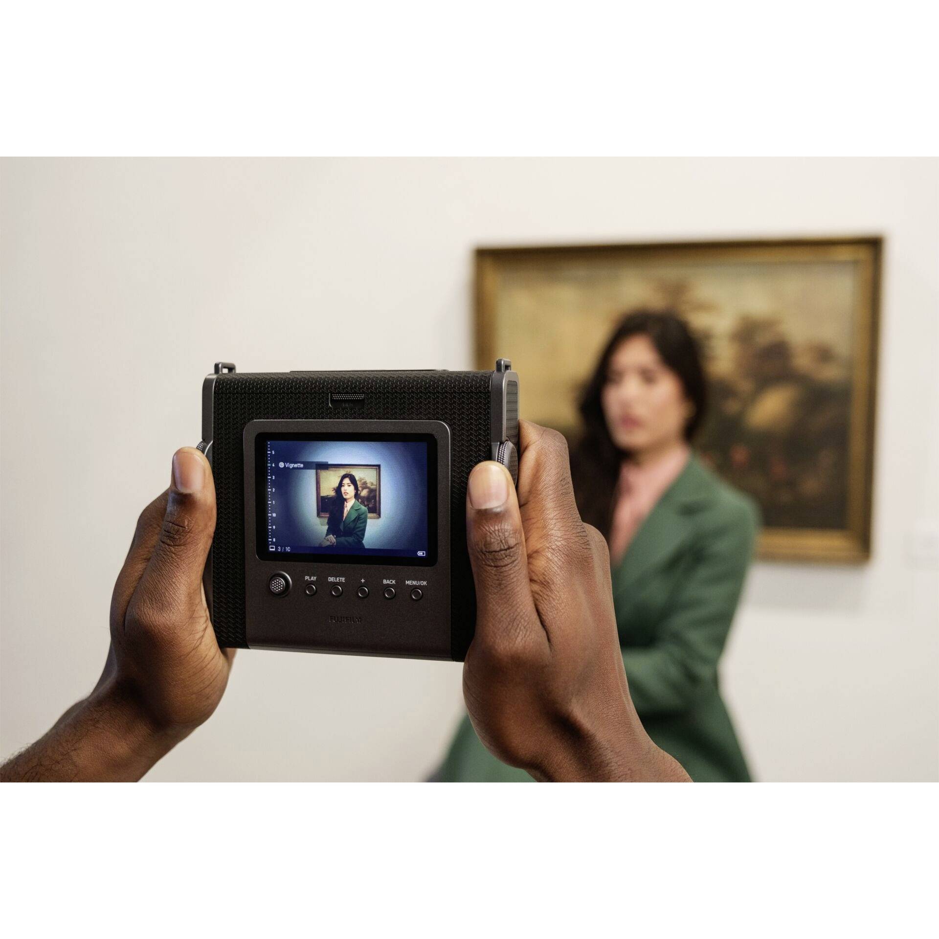 Una persona è fotografata in una stanza con un'opera d'arte sullo sfondo. Sul display della fotocamera è visibile l'immagine della persona.