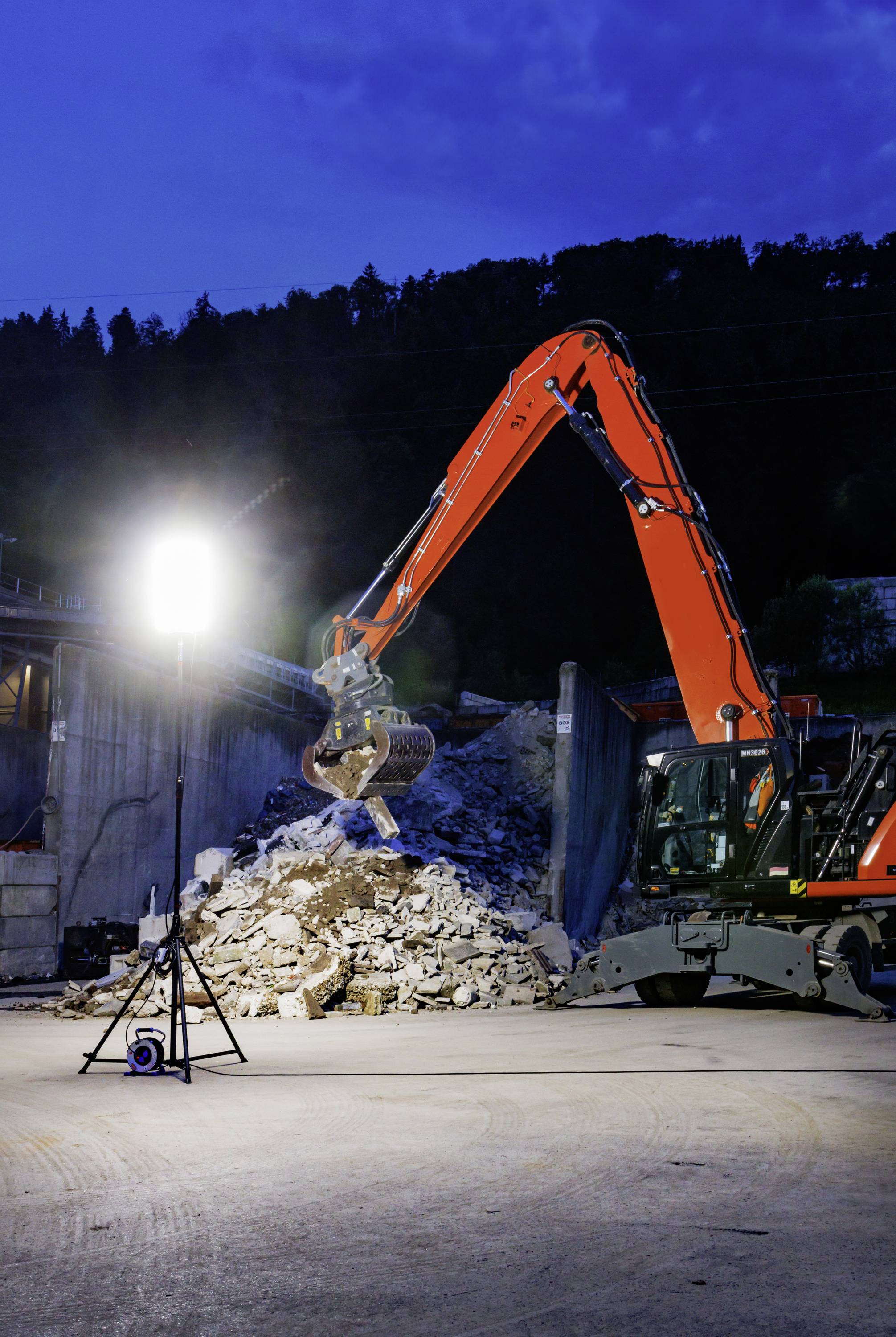 Un escavatore solleva macerie di pietra di notte in un cantiere, illuminato da una lampada brillante. Sullo sfondo si vedono alberi e un cielo blu.