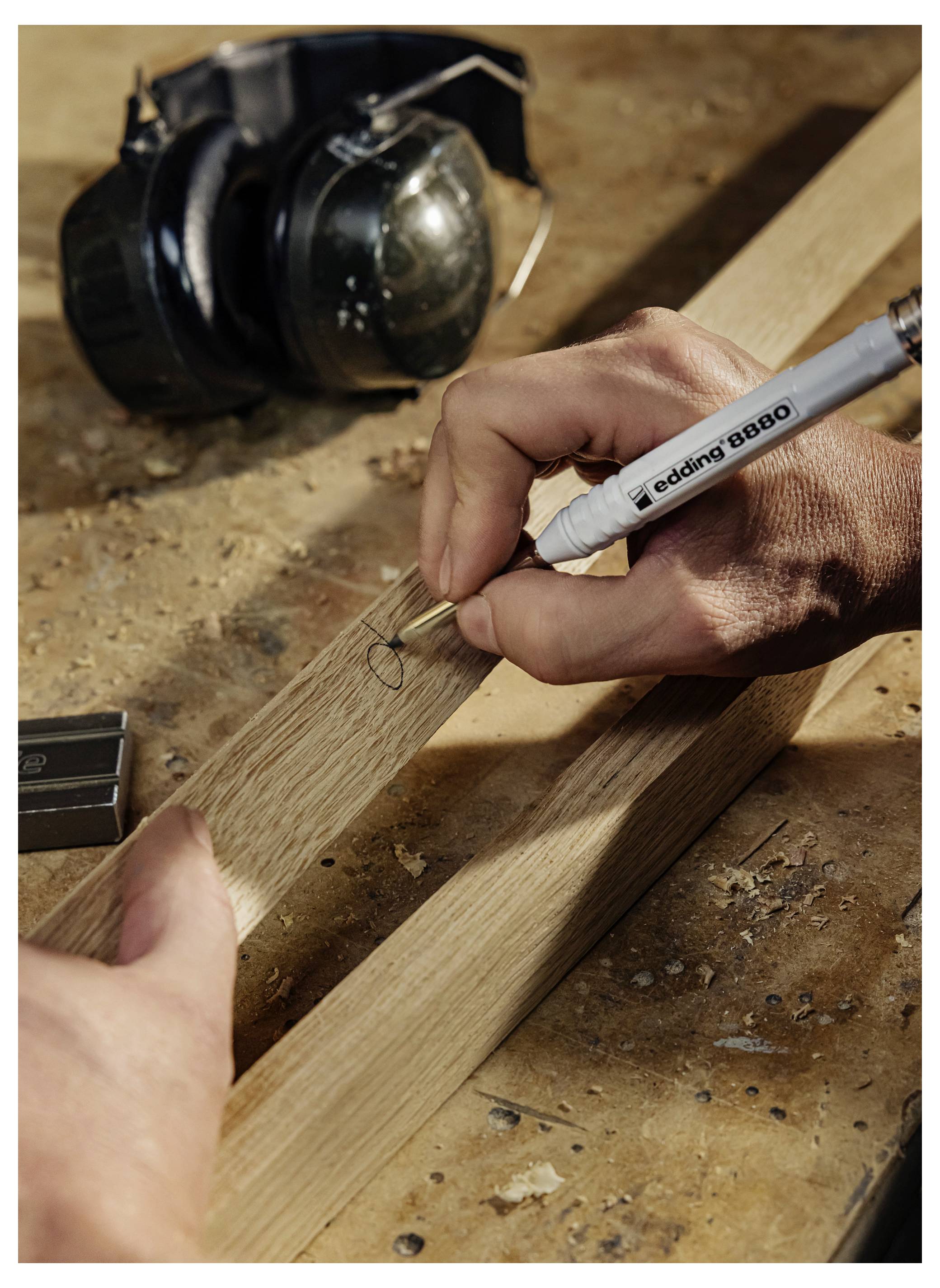 Primo piano delle mani che marcano un'asse di legno con una penna su un banco da lavoro, con protezioni acustiche per la sicurezza nelle vicinanze.