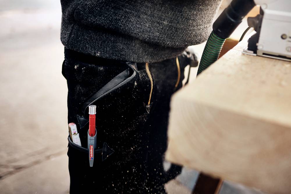 Una persona in abiti da lavoro sta tagliando legno con una sega. Una penna e un metro a nastro sono infilati in una tasca degli attrezzi agganciata alla cintura.