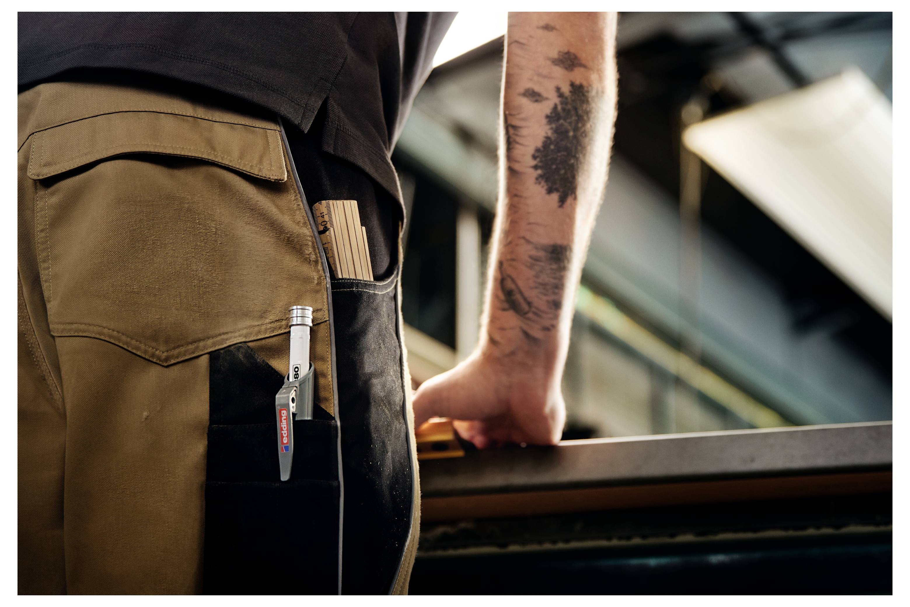 Primo piano di un operaio in pantaloni beige con una penna nella tasca, che appoggia un braccio tatuato su una ringhiera in un ambiente industriale.