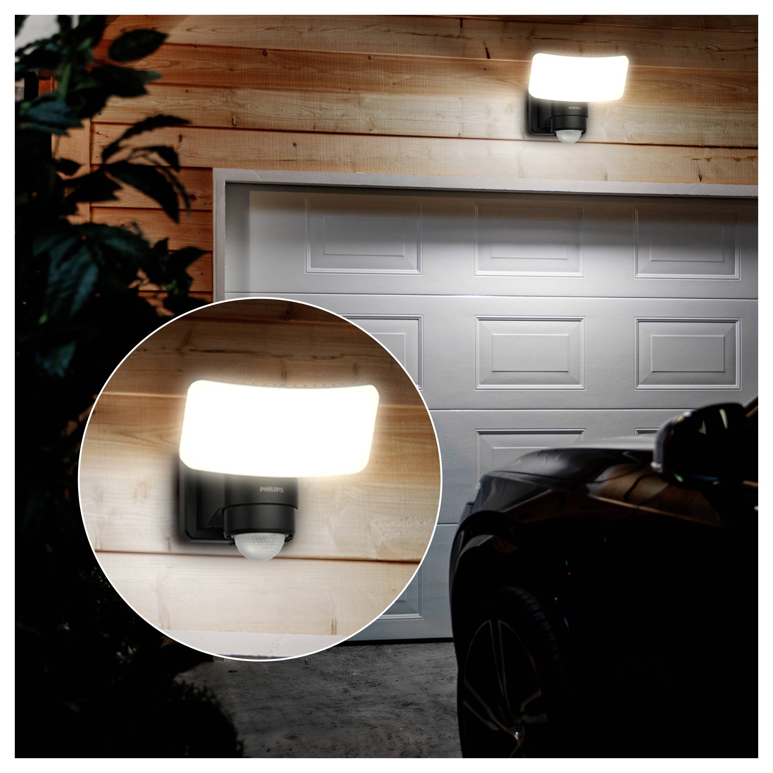 Luce con sensore di movimento montata sulla parete esterna sopra una porta di garage, mostrata accesa di notte con un ingrandimento circolare in primo piano del dispositivo.
