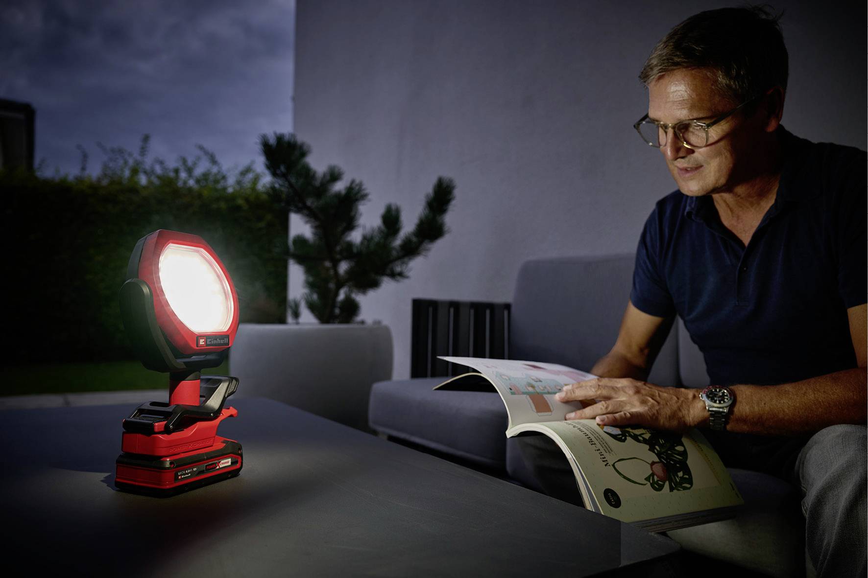 Un uomo siede in una stanza semibuia e legge una rivista, mentre una lampada accesa e luminosa su un tavolo diffonde luce.