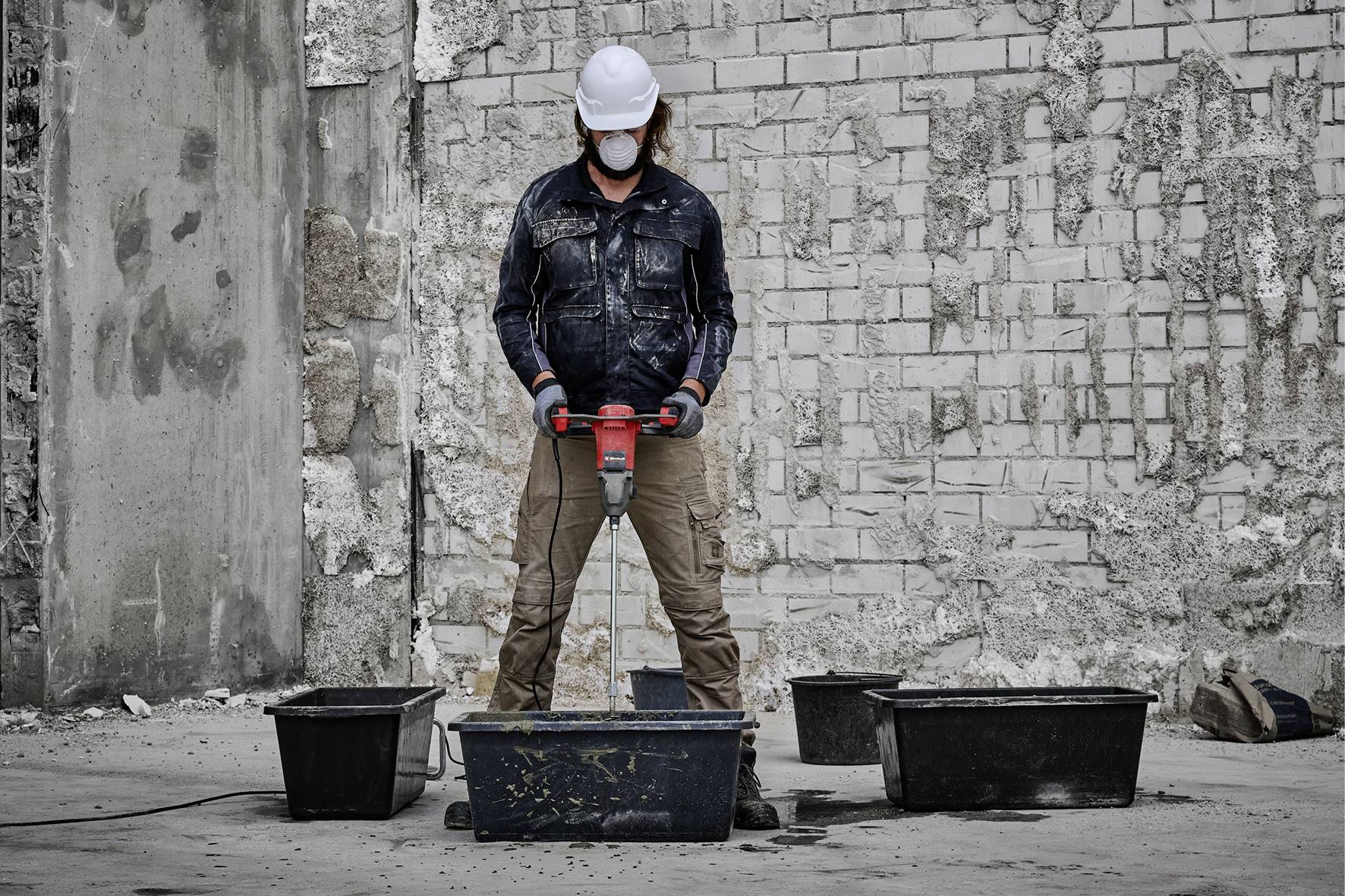 Una persona con casco protettivo e maschera sta utilizzando un martello pneumatico in un cantiere edile, circondato da diversi grandi contenitori.