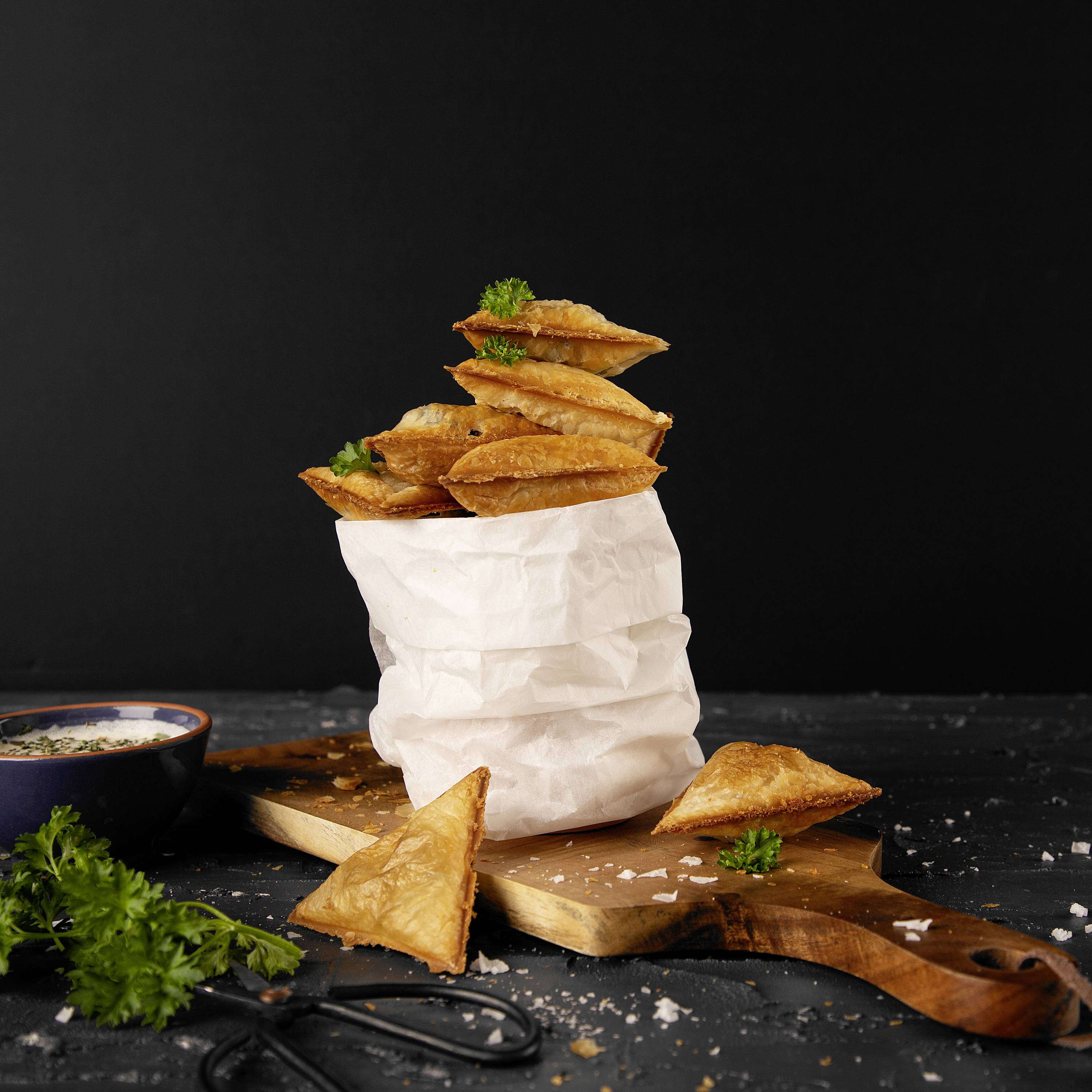 Un mucchio di dolci sfogliati su carta marrone, decorato con erbe, poggia su un tagliere. Accanto sono posizionati una ciotola con salsa e erbe fresche.
