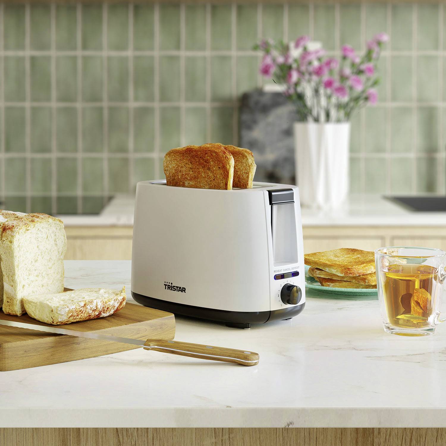 Un tostapane con fette di pane su un bancone da cucina. Accanto del pane tostato imburrato, un coltello e un bicchiere di tè. Sullo sfondo un vaso di fiori.
