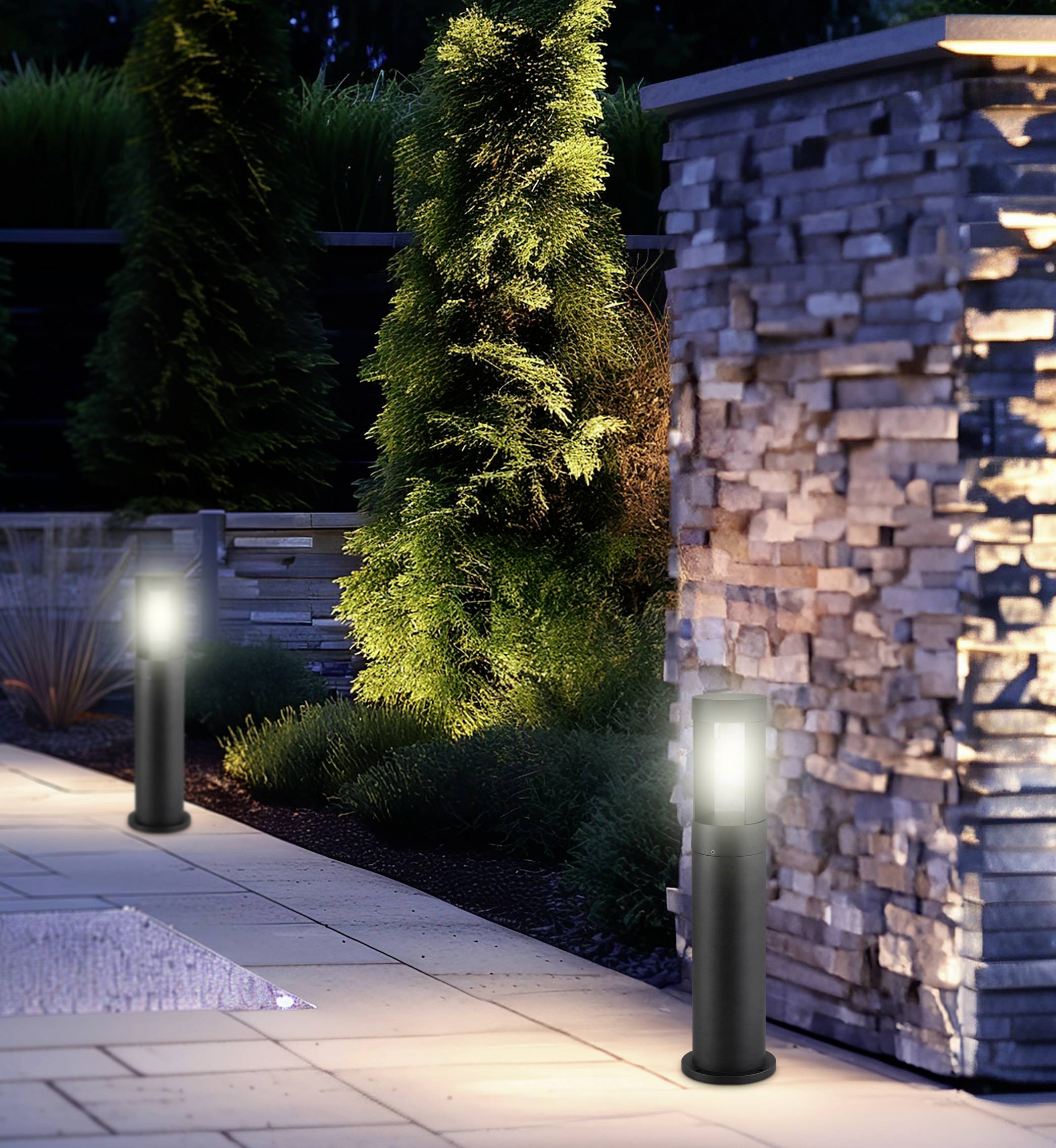 Un patio esterno è illuminato con soffusa luce da due moderni proiettori a colonna neri. Sullo sfondo sono visibili un muro in pietra e cespugli alti.