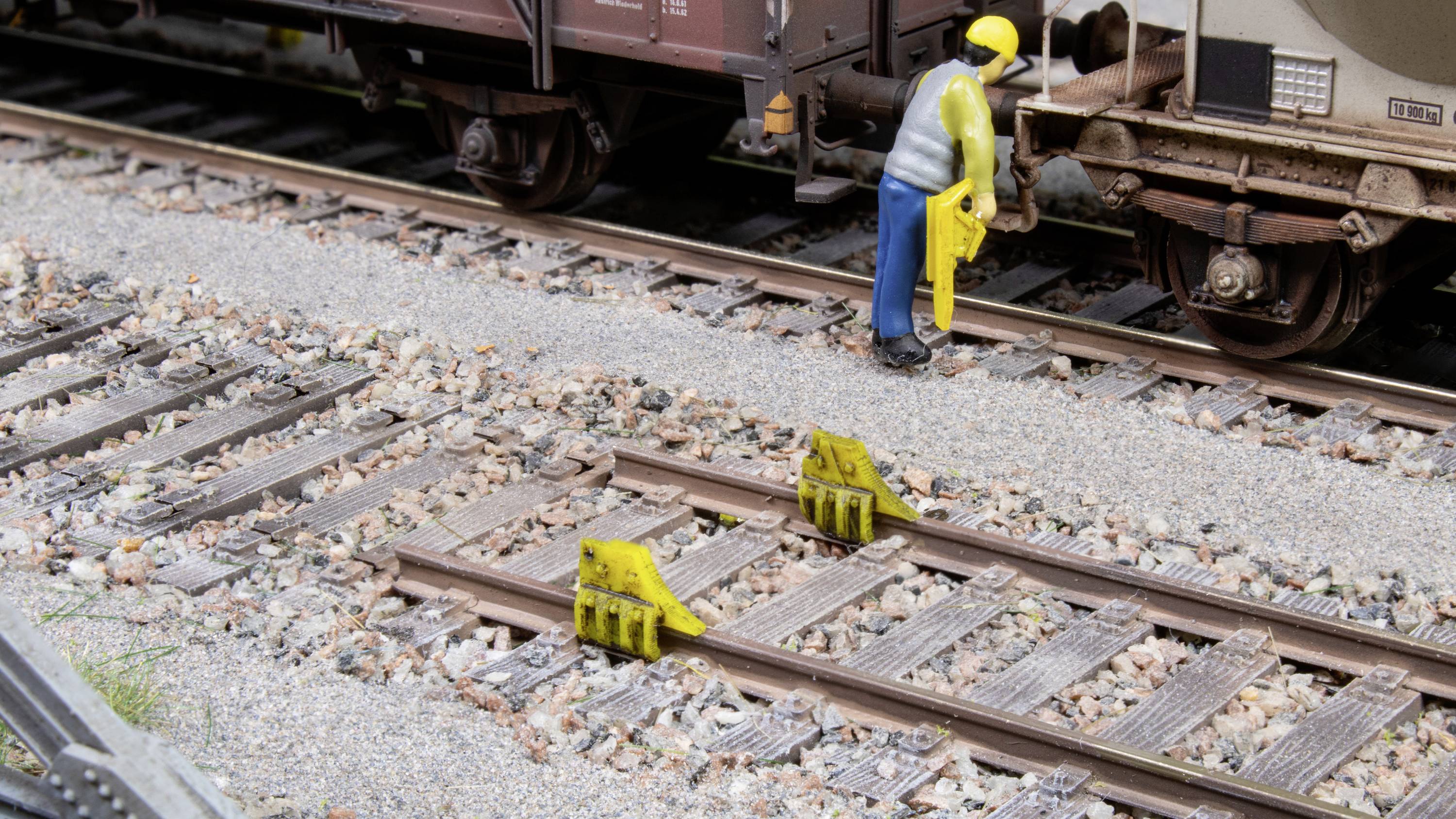 Un lavoratore ferroviario in miniatura è in piedi tra i binari e tiene in mano un attrezzo giallo. I dispositivi di delimitazione dei binari sono visibili sulle rotaie.