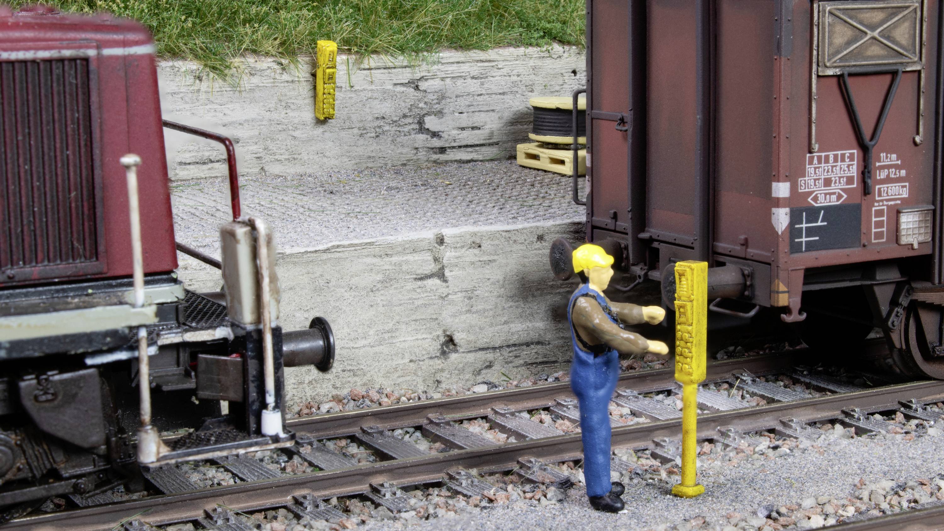 Un addetto al modellismo ferroviario con tuta blu e casco giallo sta azionando un segnale giallo sulla linea ferroviaria accanto a un vagone merci.