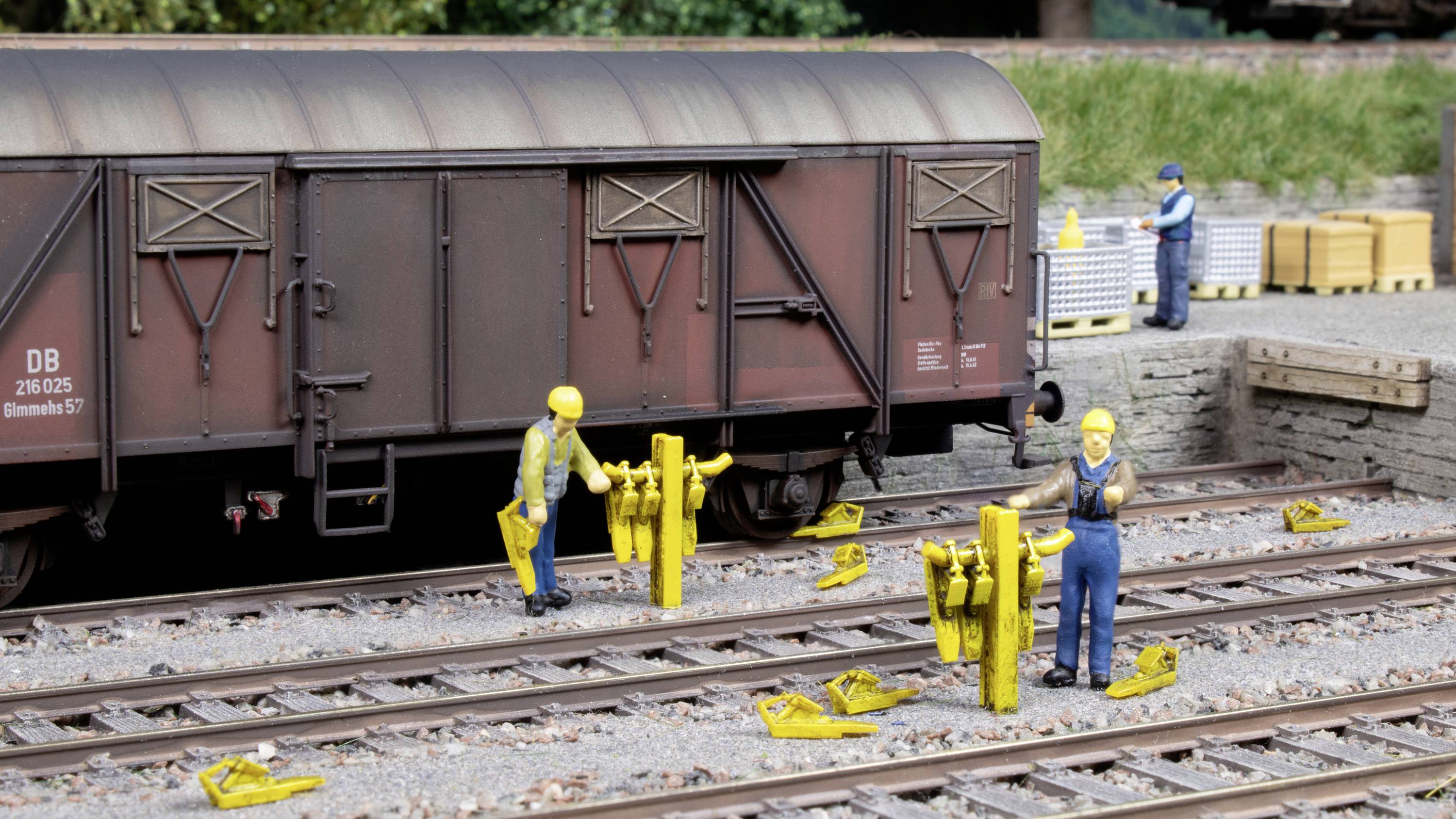 Una scena di un plastico ferroviario mostra operai che lavorano con attrezzi gialli sui binari ferroviari, mentre un vagone merci è sullo sfondo.