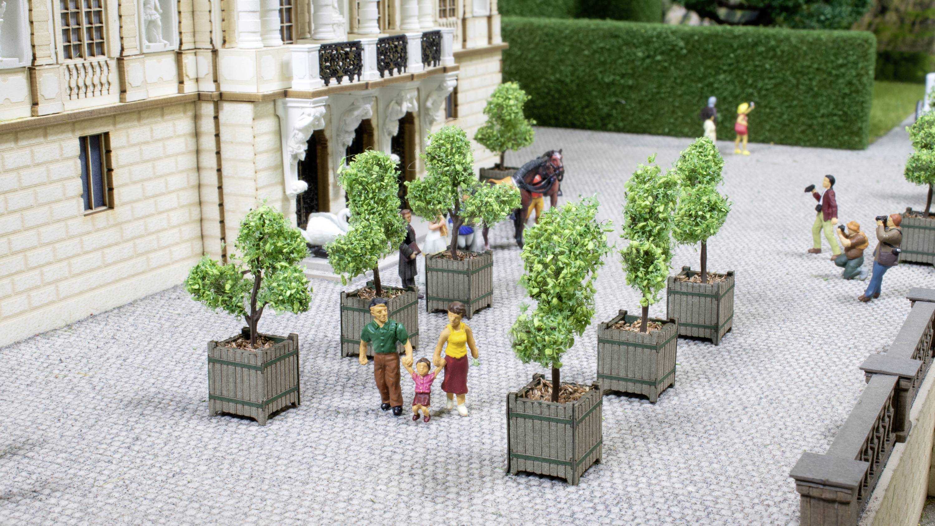 Un modellino mostra persone che camminano in un cortile lastricato tra alberi in vaso. Una coppia tiene per mano un bambino.
