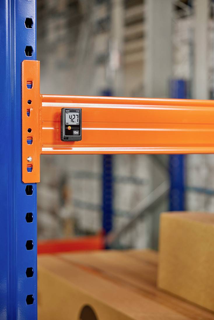 Termometro su uno scaffale arancione in un magazzino. Il display digitale indica 42,7 gradi. Sullo sfondo, scatole sfocate.