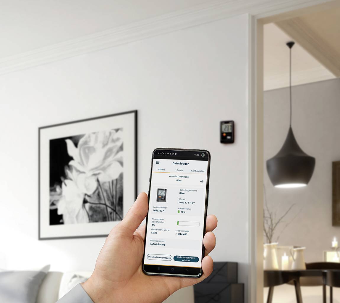 Una persona tiene uno smartphone con un'app di controllo del riscaldamento aperta; sullo sfondo un soggiorno moderno con illuminazione elegante.