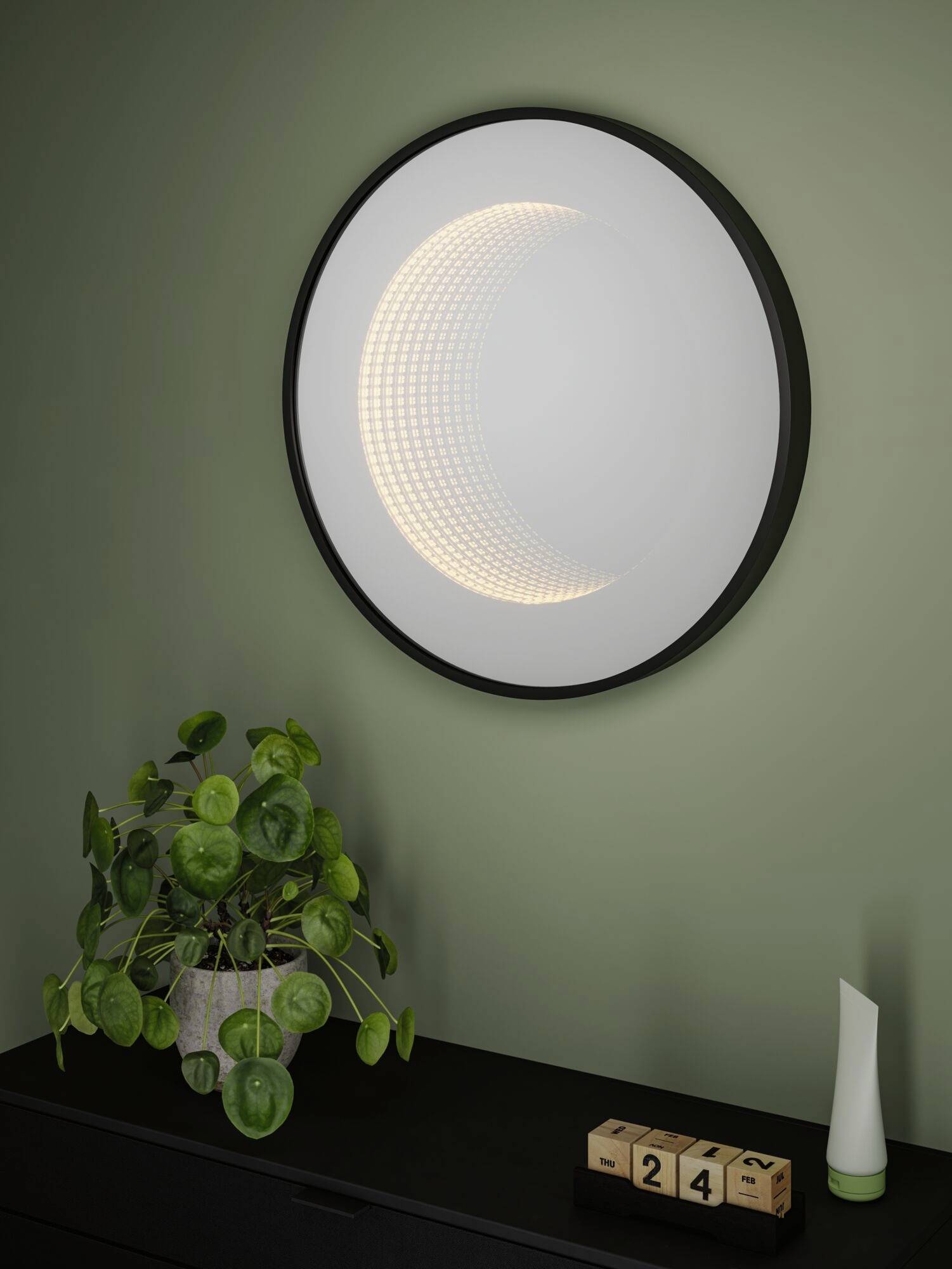 Uno specchio rotondo con illuminazione a mezzaluna è appeso a una parete verde. Sotto di esso c'è un tavolo con una pianta e un calendario.