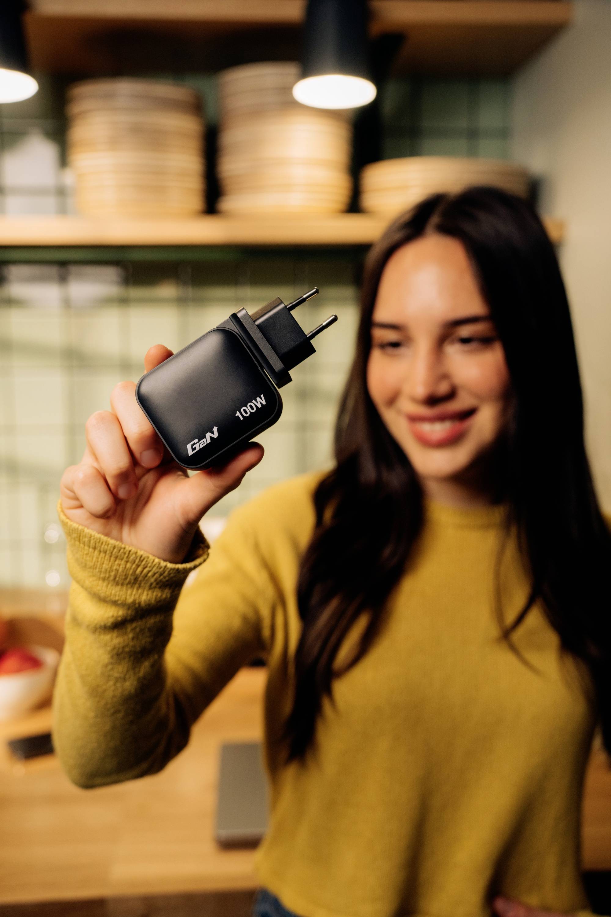 Una donna tiene in mano un caricabatterie con la scritta '100W' e sorride. Sullo sfondo sono visibili elementi di cucina.