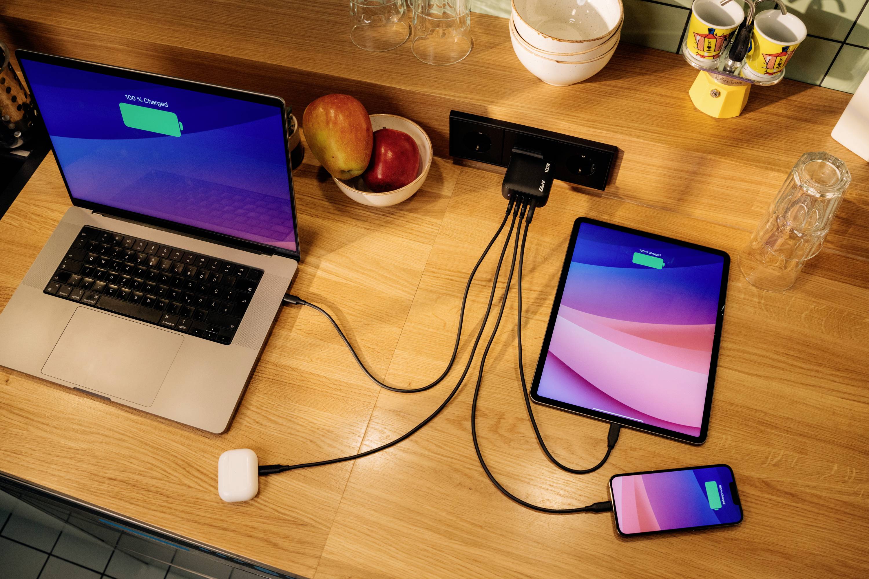 Un laptop, un tablet e uno smartphone vengono caricati su un tavolo di legno tramite una presa multipla, accanto a una ciotola di frutta.