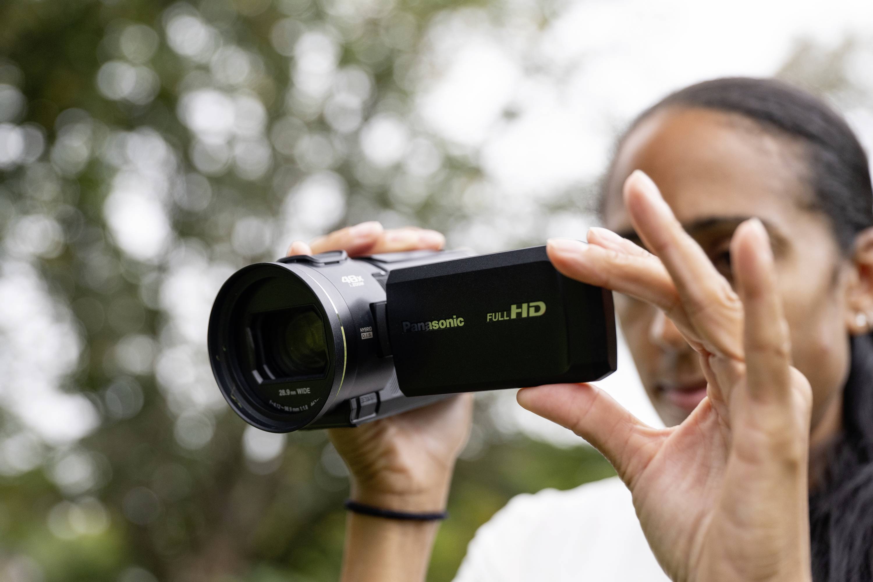 Una persona tiene una videocamera nera all'aperto, concentrata sull'obiettivo. Sullo sfondo si intravedono alberi sfocati.