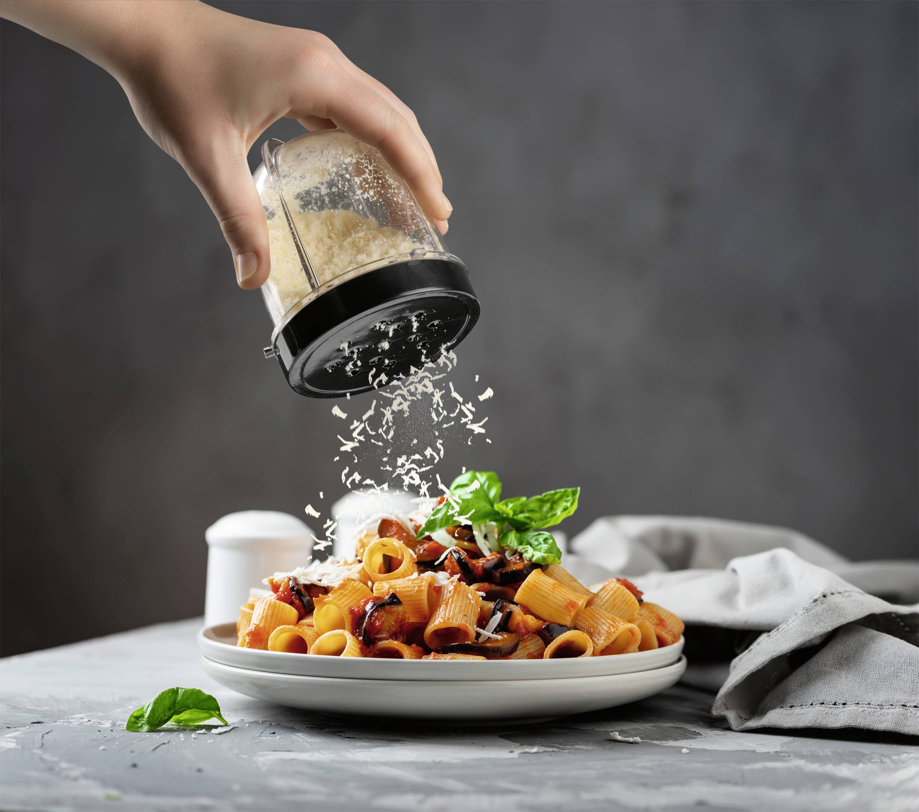 Una mano sparge formaggio grattugiato su un piatto di pasta con sugo di pomodoro, guarnito con basilico. Sfondo sfocato.