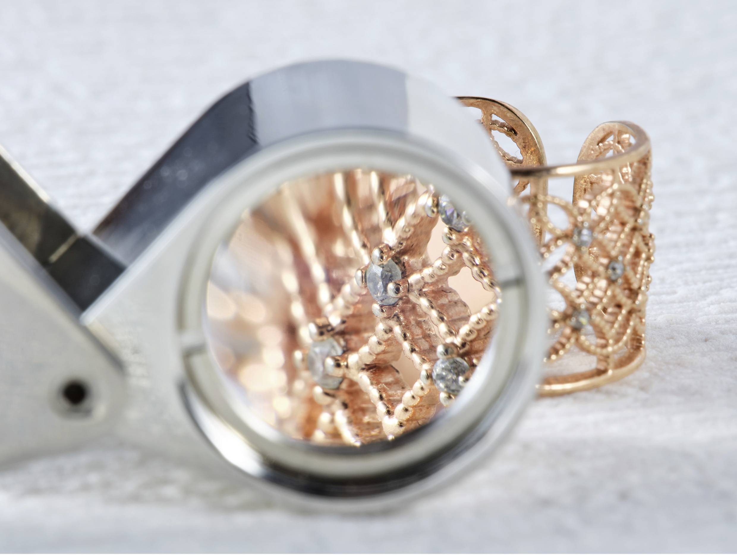 Una lente di ingrandimento si concentra su un anello intricato in oro con decorazioni di diamanti, evidenziando la lavorazione dettagliata su uno sfondo testurizzato.