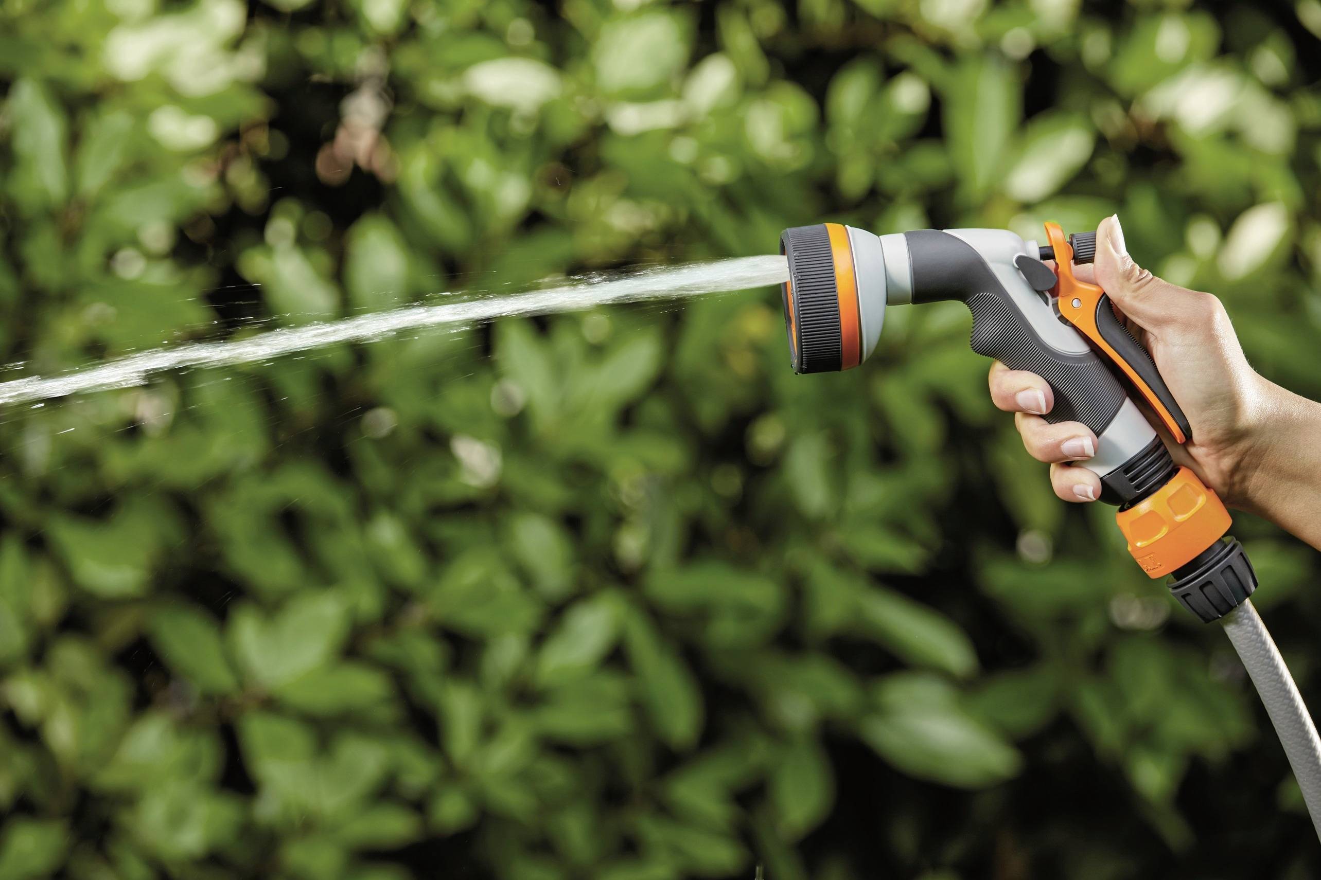 Una mano regge un tubo da giardino con ugello a spruzzo, da cui scorre acqua, sullo sfondo foglie verdi sfocate.