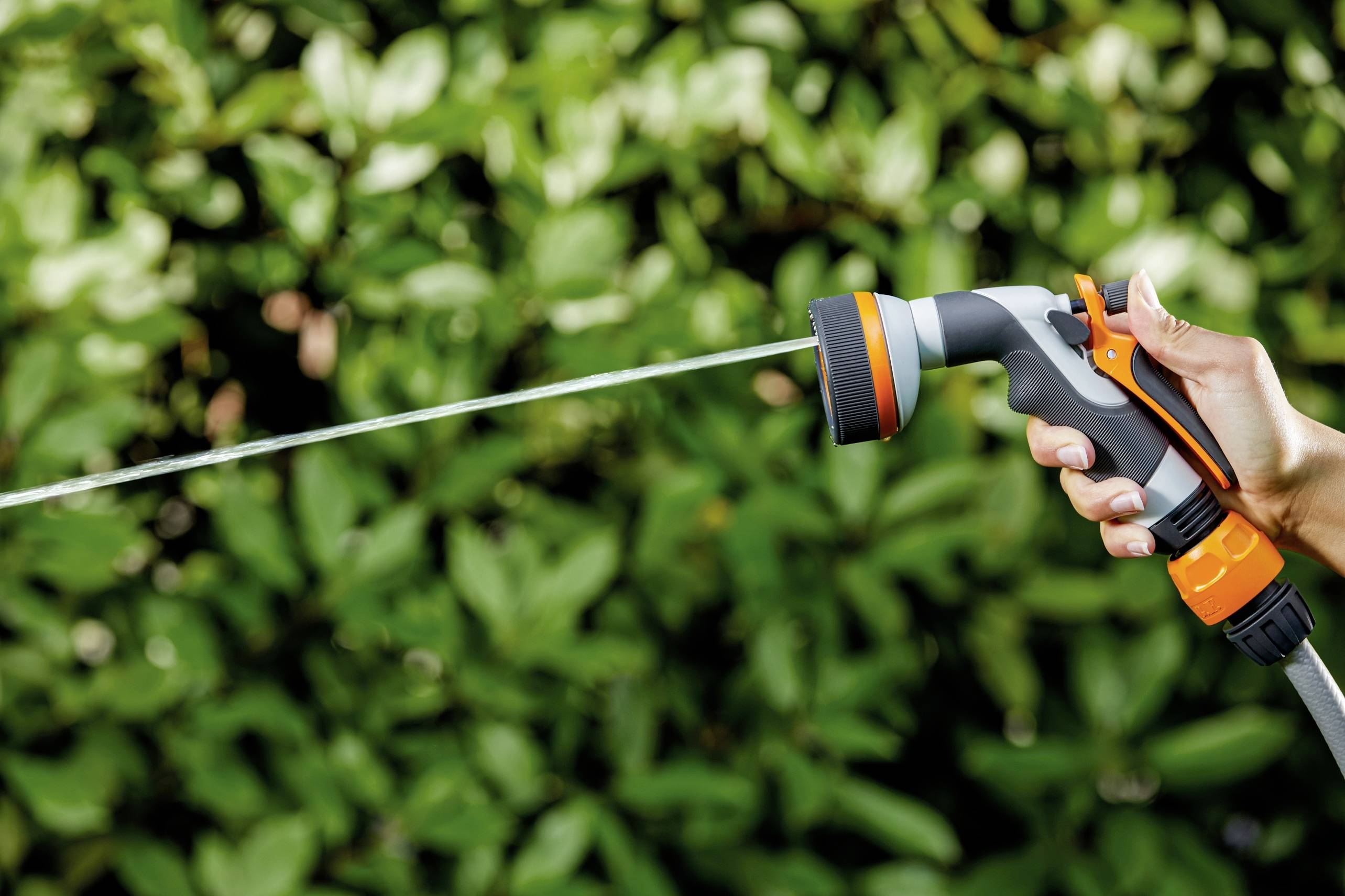 Una mano tiene un nebulizzatore da giardino da cui un getto d'acqua spruzza su foglie verdi sullo sfondo.
