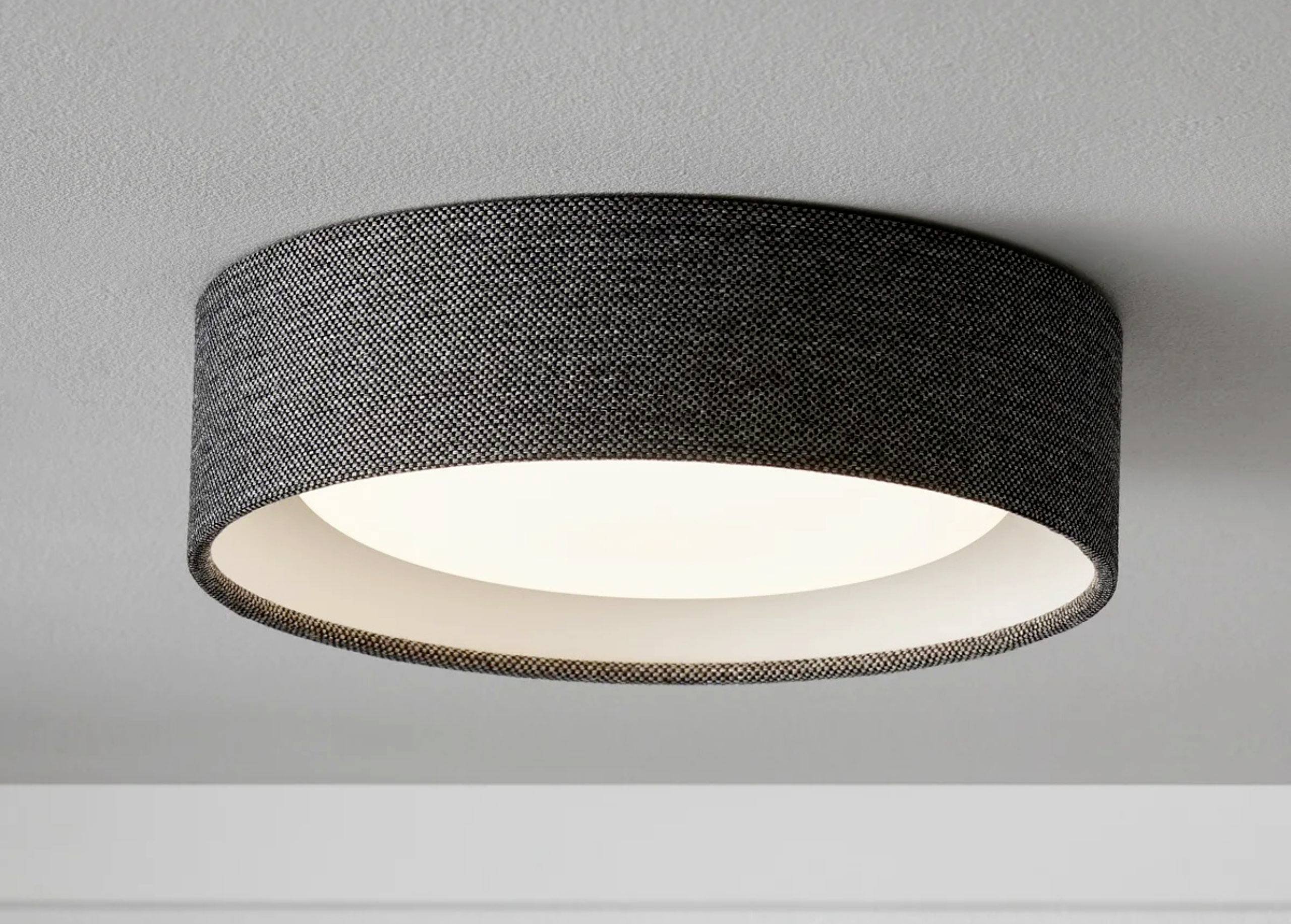Un'applique rotonda con rivestimento in tessuto nero che emana una luce calda. Montata su un soffitto bianco.