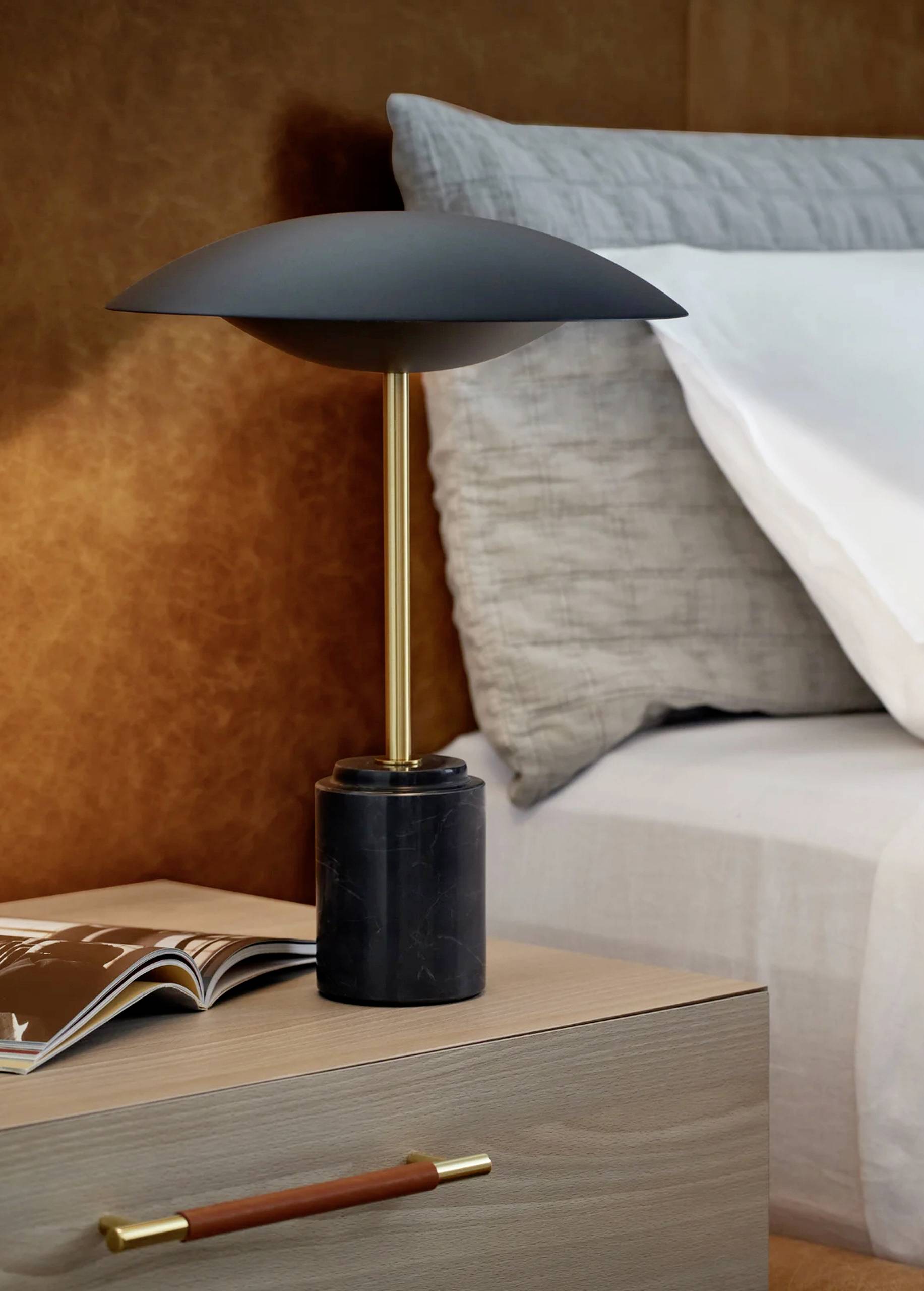Lampada da tavolo moderna con paralume nero e base dorata su un comodino accanto a un libro aperto, con un letto sullo sfondo.
