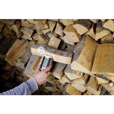 Una persona misura l'umidità di un pezzo di legno con un misuratore di umidità davanti a una pila di legna da ardere.