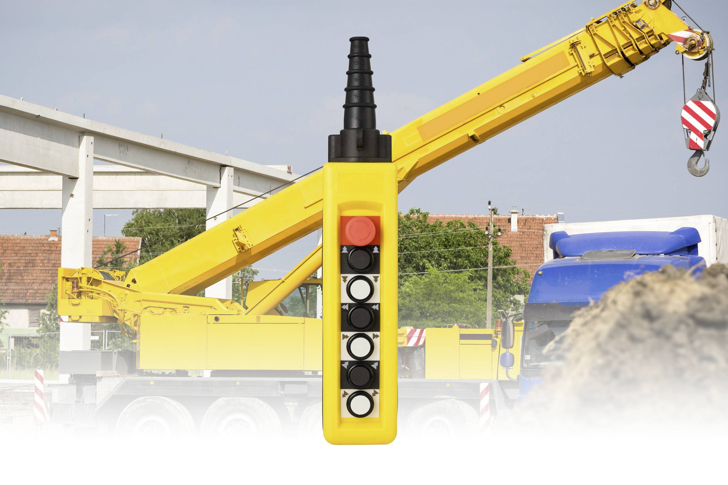 Un dispositivo di telecomando giallo è in primo piano, con una gru da cantiere che solleva materiali sullo sfondo di un cantiere.